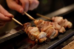 two hands holding chopsticks over some meat on a grill with another hand reaching for the skewer