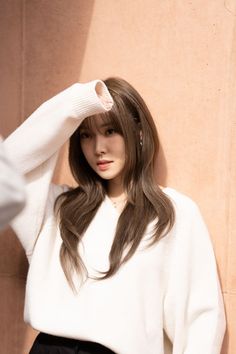 a woman with long hair standing next to a wall wearing a white sweater and black pants