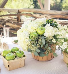 an arrangement of flowers and apples in a basket on top of a white table cloth