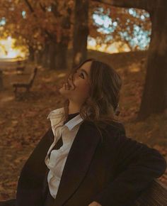 a woman sitting on top of a wooden bench in a park next to trees and leaves