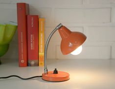 an orange desk lamp sitting on top of a white table next to books and a green vase