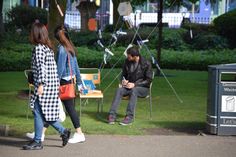 two women and a man are sitting on chairs in the park, one is looking at his cell phone