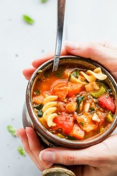 two hands holding a bowl of pasta and vegetable soup with a spoon in the bowl