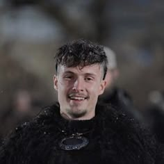 a man in a black fur coat smiles at the camera while people watch from behind him