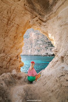 a woman sitting on the edge of a cave looking out at the water and mountains