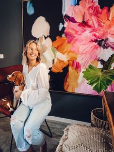 a woman sitting on a chair holding a dog in front of a large flower painting