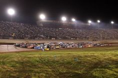 many cars are lined up on the dirt track at night in front of an empty stadium