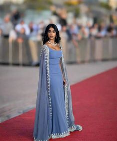 a woman in a blue gown and cape standing on a red carpet with people behind her