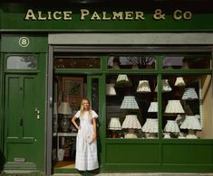 a woman standing in front of a green storefront with lamps on the outside and inside