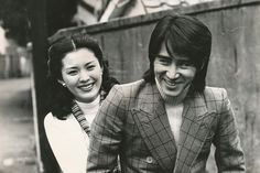 a man and woman standing next to each other in front of a wooden fence smiling at the camera