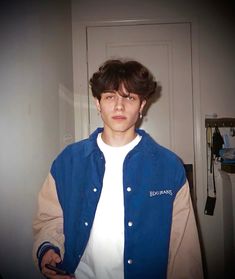 a young man standing in front of a door wearing a blue jacket and white t - shirt