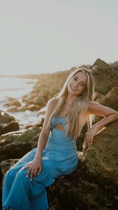 a woman in a blue dress is sitting on rocks by the ocean with her legs crossed