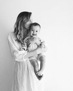 a woman holding a baby in her arms and smiling at the camera while standing against a white wall