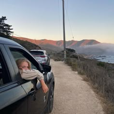 a woman leaning out the window of a car on a dirt road next to the ocean