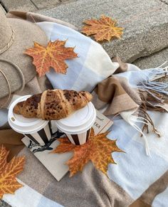 a coffee cup and croissant on a blanket with autumn leaves around it,