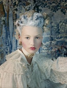 a woman with blue hair wearing a white dress and gold earrings in front of a painting