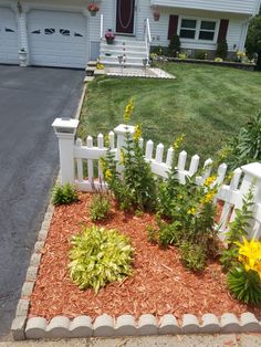 a white picket fence sitting in front of a house next to a flower bed with yellow flowers