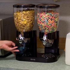 two machines filled with different kinds of cereal on top of a counter next to a microwave