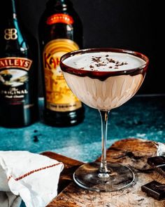 a drink in a coupe glass on a cutting board next to two bottles of booze