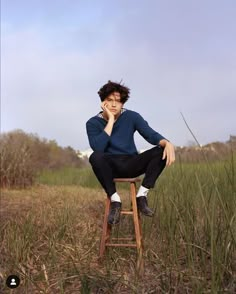 a young man sitting on top of a wooden chair in a field with tall grass