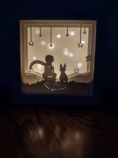 an illuminated shadow box with a boy and two cats sitting on the ground in front of it