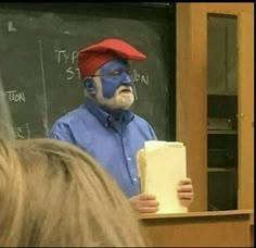 a man with blue face paint holding a piece of paper in front of a blackboard