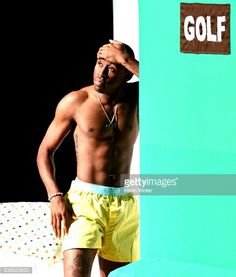 a shirtless man in yellow shorts leaning against a blue wall with the word golf on it