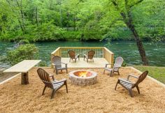 a fire pit surrounded by lawn chairs and tables in front of a river with trees