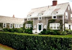 a large gray house with white trim and windows next to a lush green hedge in front of it