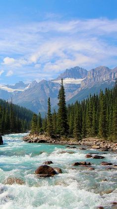 a river with rapids and rocks in the middle surrounded by pine trees, mountains and snow capped peaks