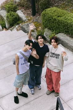 three young men standing next to each other in front of some rocks and bushes with their hands up