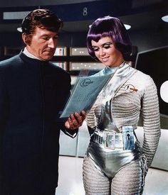 the man and woman are dressed in futuristic garb, with one holding a clipboard