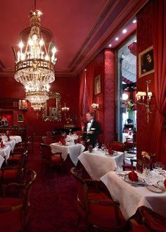 a restaurant with red walls and chandeliers hanging from the ceiling, tables set for dinner