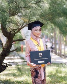 a woman wearing a graduation gown and holding a plaque in front of a pine tree