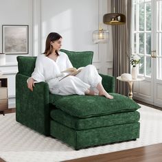 a woman sitting on a green chair reading a book in her living room with a large window