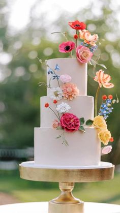 a three tiered cake with flowers on top is sitting on a gold stand outside