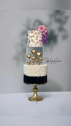 a three tiered cake with gold hearts and flowers on the top is decorated in white, black, and pink