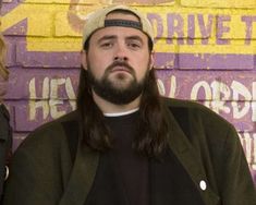 a man with long hair and beard standing in front of a brick wall wearing a hat