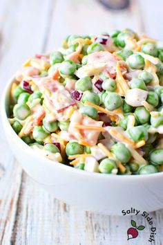 a white bowl filled with peas and cheese on top of a wooden table next to an apple