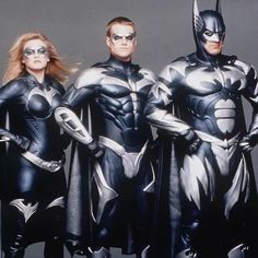 three people dressed as batman and catwoman standing in front of a gray background with their hands on their hips