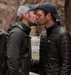 two men standing next to each other in front of a mountain river and one is kissing the other