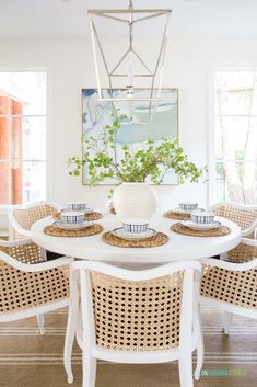 a dining room table with wicker chairs and plates on it, in front of a painting