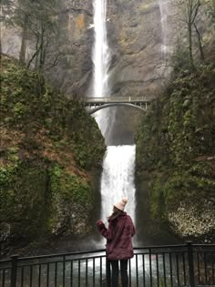 a woman standing in front of a waterfall with a bridge over it and trees around her