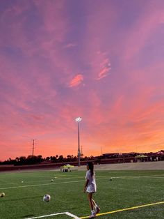 a woman standing on top of a lush green field next to a soccer ball under a pink sky