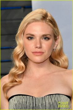 a blonde woman with wavy hair wearing a strapless dress and diamond earrings, looking at the camera