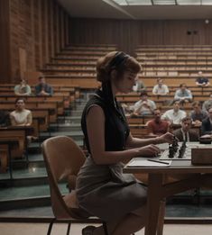 a woman sitting at a table playing chess with other people in the auditorium behind her