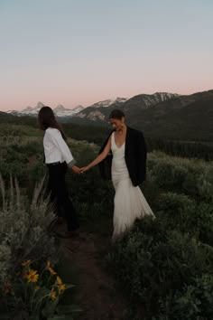 a bride and groom holding hands walking through the mountains at sunset in their wedding attire