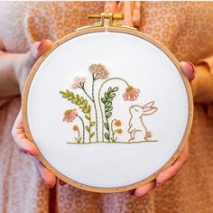 a person holding a small embroidery project with flowers and a bunny in the grass on it