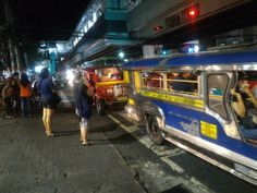 people are standing on the side walk next to parked cars and buses at night time