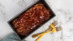 a pan filled with meat covered in sauce next to two yellow utensils on top of a marble counter